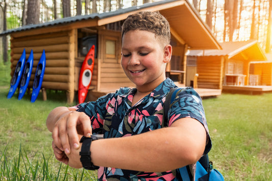 Smiling boy at summer camp checking GPS smartwatch in front of log cabin with kayaks — myFirst Fone R2 keeps kids connected and safe outdoors