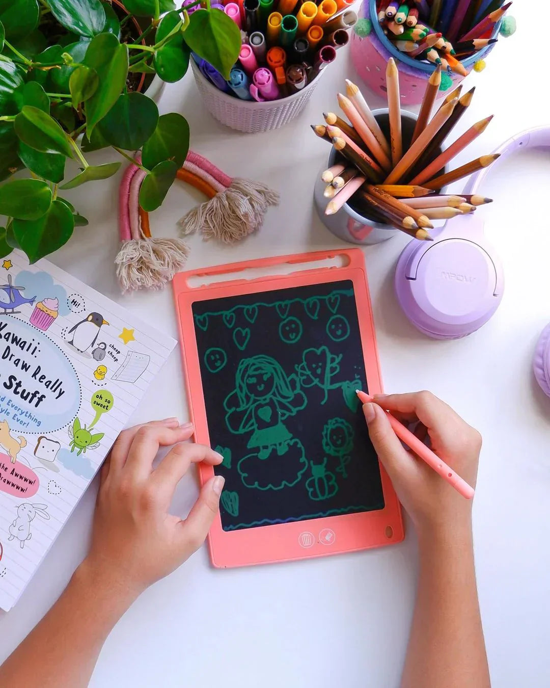 Child using a pink myFirst LCD writing tablet for kids to draw sea creatures, surrounded by pencils, markers, and creative art supplies on a white desk.