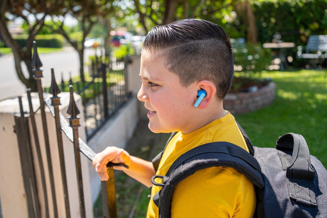 A young boy wearing myFirst wireless earbuds walks outdoors with a backpack on, smiling and engaged. The child-safe earbuds fit snugly, ideal for on-the-go listening.
