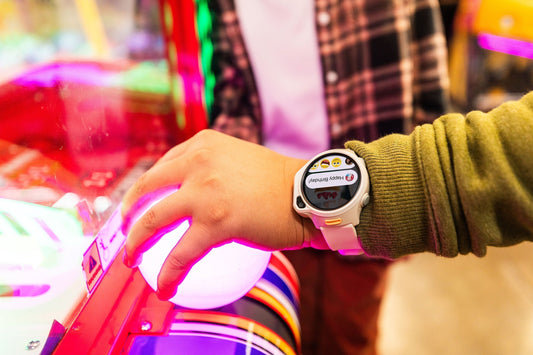 Boy's hand closeup at arcade wearing kids smart watch with gps and video call screen showing Happy Birthday text message, with camera and simple buttons showing ease of use for young children.