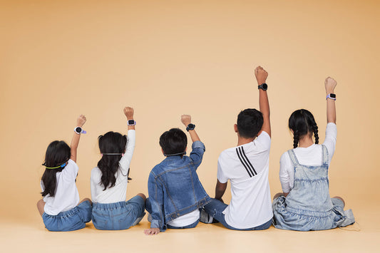 Group of kids sitting together, raising their arms to show colorful kids’ smartwatches with SIM card, video calling, GPS tracking, and distraction-free school mode – an alternative to smartphones for safe communication.