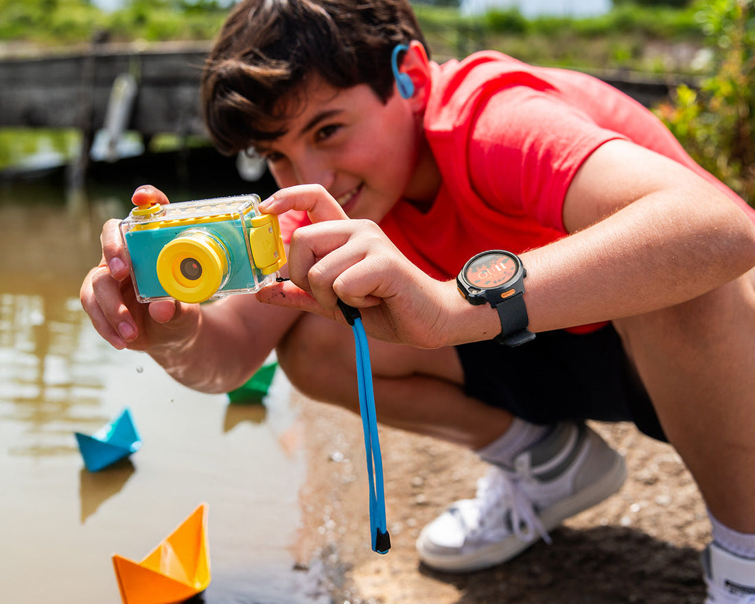 Boy wearing a kids’ GPS smartwatch and open-ear headphones takes photos with a waterproof digital camera near a lake, surrounded by colorful paper boats.