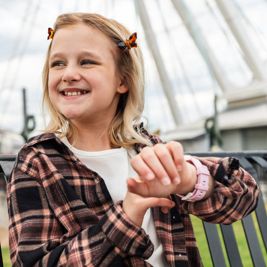 Smiling young girl wearing butterfly hair clips and a plaid shirt, checking her pink myFirst Fone R2 smartwatch with GPS and video call features, seated outdoors near a large Ferris wheel.