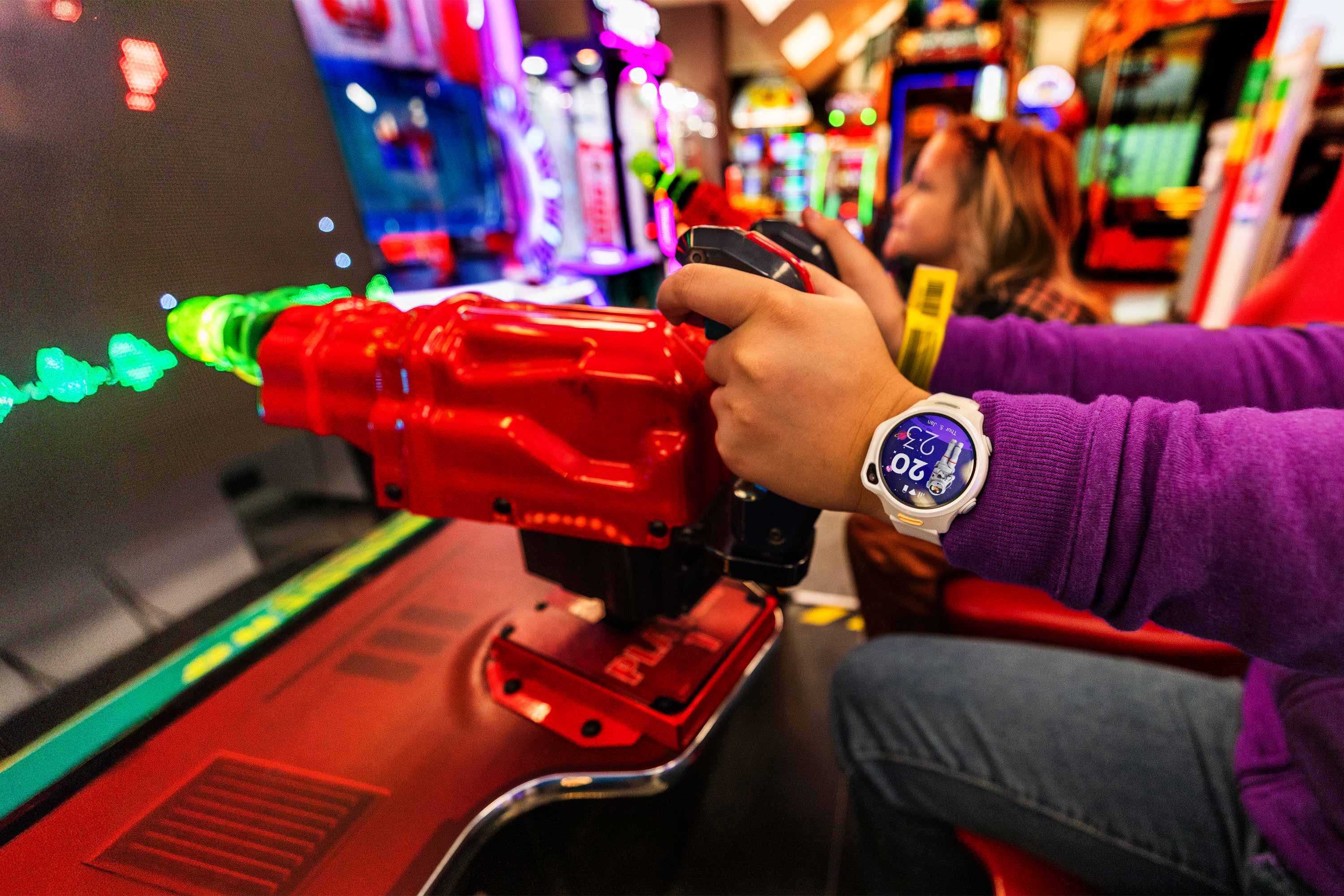 Child wearing the myFirst Fone R2 smartwatch while playing an arcade game. The smartwatch includes GPS tracking, video calling, text messaging, a fitness tracker, and a parental control app. It has no social media, no third-party apps or games—designed for safety, connection, and kid-friendly independence.