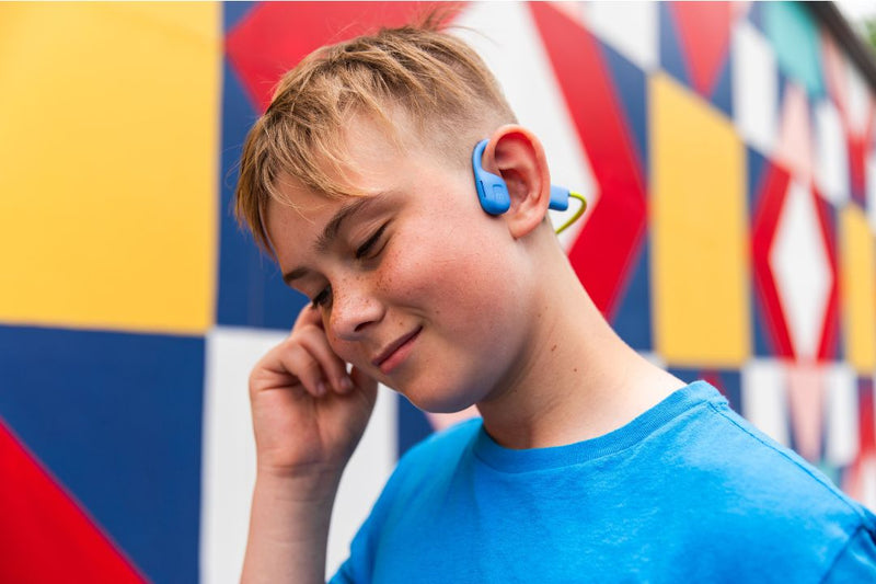 A young boy in a blue shirt outside is wearing Kids Headphones that don't cover his ears, keeping them free so he can hear the environment.