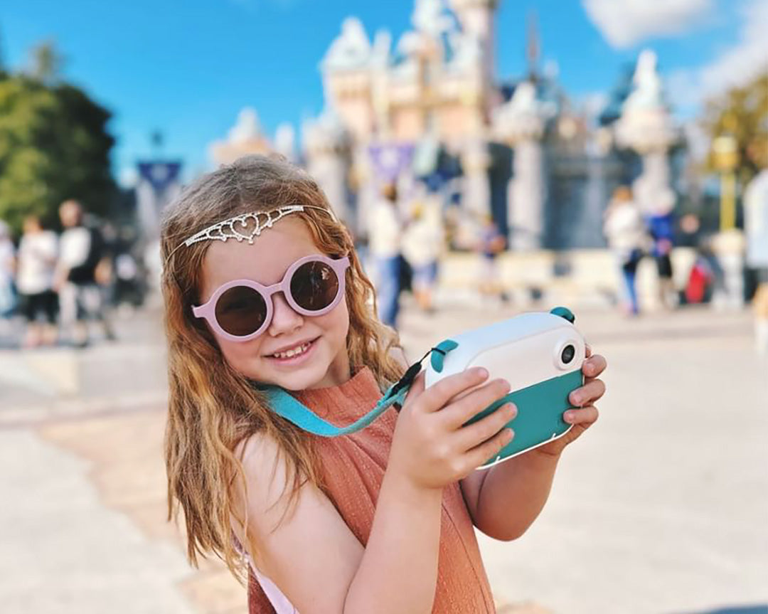 Young girl wearing sunglasses and tiara using instant print kids camera at theme park, demonstrating myFirst Camera Insta 2 for travel journaling