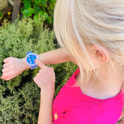 myFirst Fone R2 kids smartwatch worn by a young girl in a pink top, pointing at the watch outdoors in a garden setting.