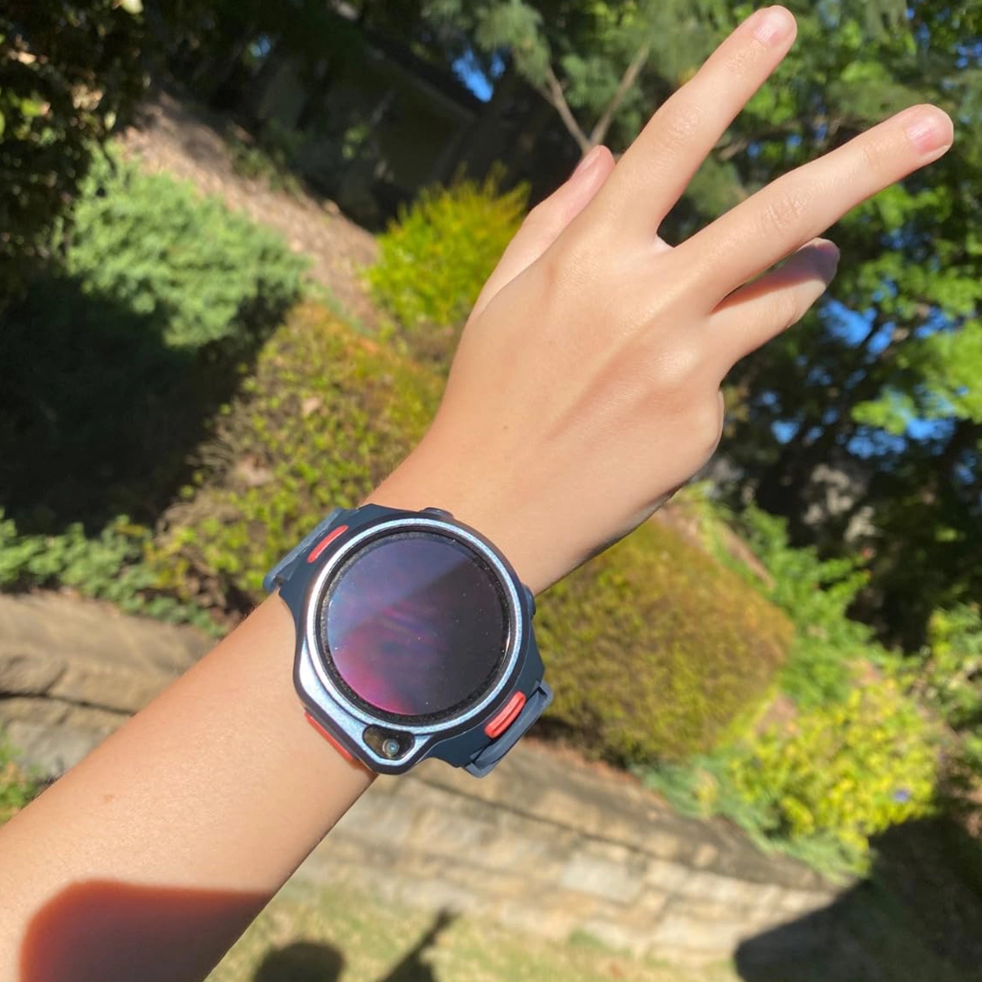 myFirst Fone R2 dark navy kids smartwatch worn on a hand making a peace sign in a sunny outdoor garden setting.