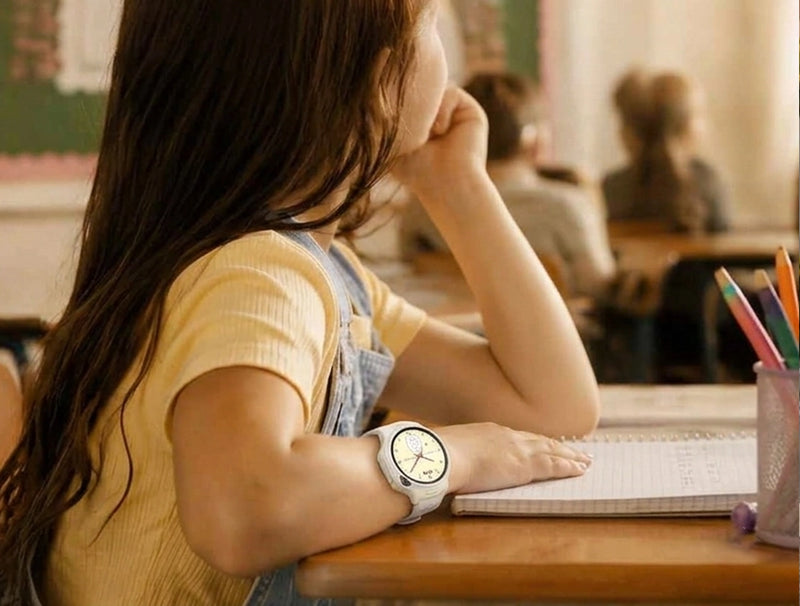 yFirst Fone R2 white kids smartwatch worn by a girl focusing on her notebook in a classroom setting, displaying a playful cartoon watch face