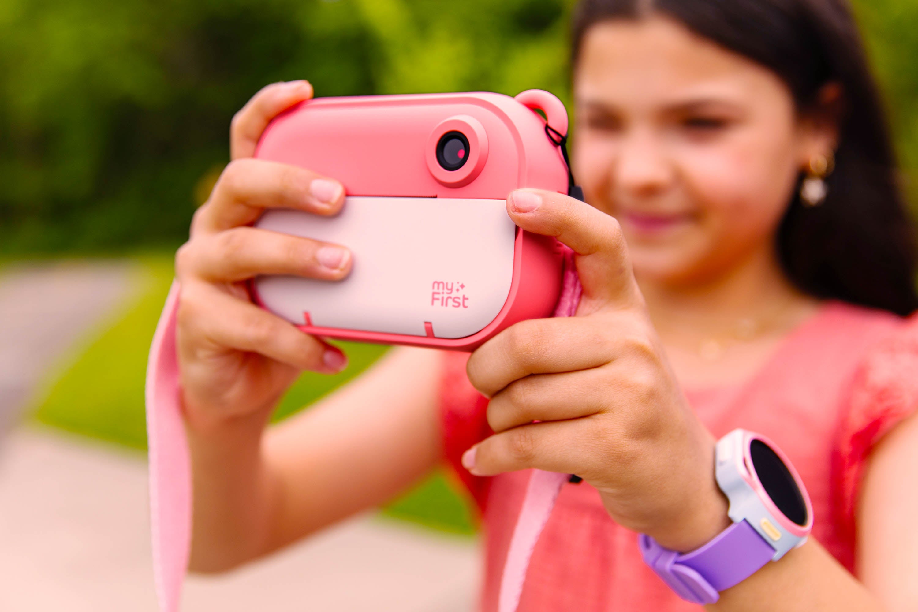 Young girl using the pink myFirst Camera Insta 2 to take a photo outdoors, wearing a kids smartwatch on her wrist. The instant print camera features 5MP resolution, thermal printing, and kid-friendly design—perfect for fun, screen-free creativity.